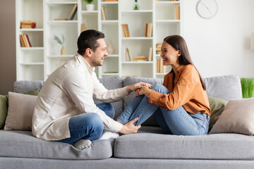 Excited man and woman sitting on couch facing each other and engaged in pleasant conversation, side view