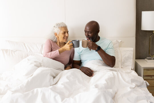 Clinking mugs Diverse senior couple leaning against pillows in bed, with modern lamp and books