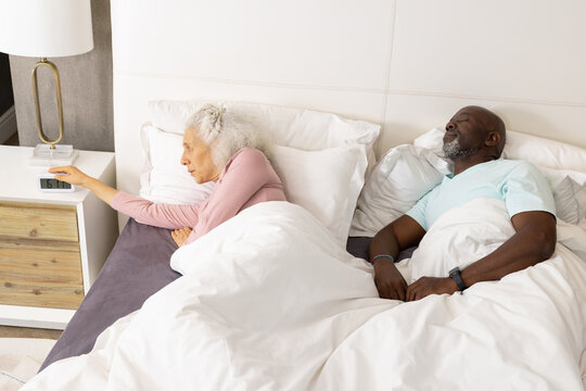 Diverse senior couple lying in bed in modern bedroom, with woman reaching digital alarm clock