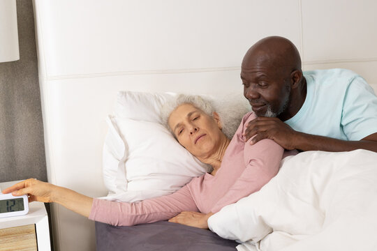 Reaching Diverse senior couple turning off digital alarm clock on wooden nightstand in bedroom