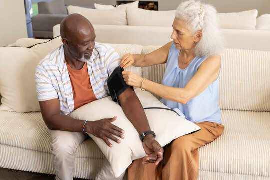 Checking diverse senior couple fitting blood pressure cuff on beige sofa at home, with throw pillow