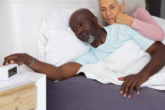 Diverse senior couple reaching for alarm clock on wooden nightstand lying in bed, with duvet