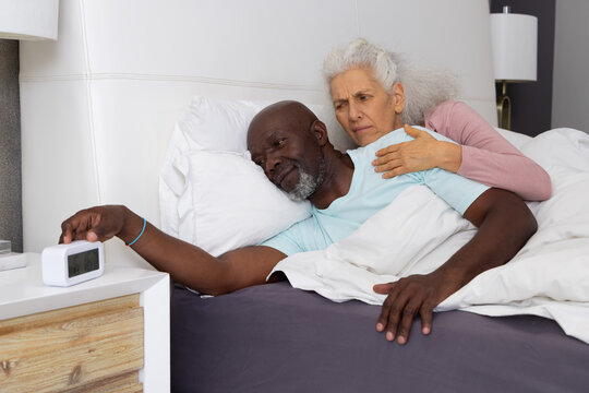 Diverse senior couple reaching for digital alarm clock while lying under white linens in bedroom