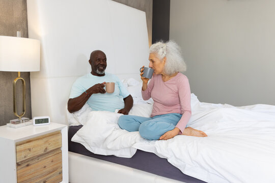 Chatting Diverse senior couple sitting on bed in modern bedroom, holding beige and gray mugs
