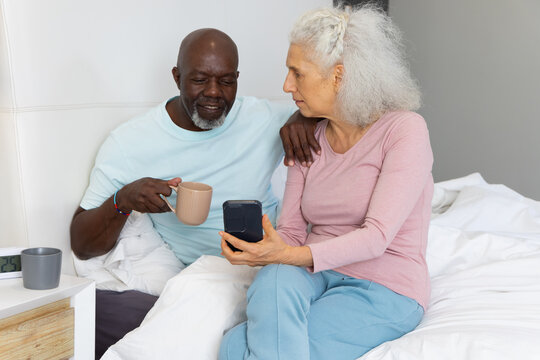 Sipping coffee senior couple sitting on bed in bedroom, holding device near digital alarm clock