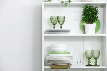 Clean glasses, plates and green houseplant on shelves near white wall indoors, space for text