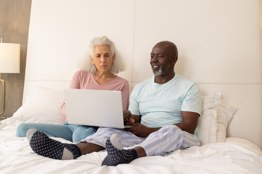 Diverse senior couple sitting on bed using silver laptop in bright bedroom, with gold floor lamp