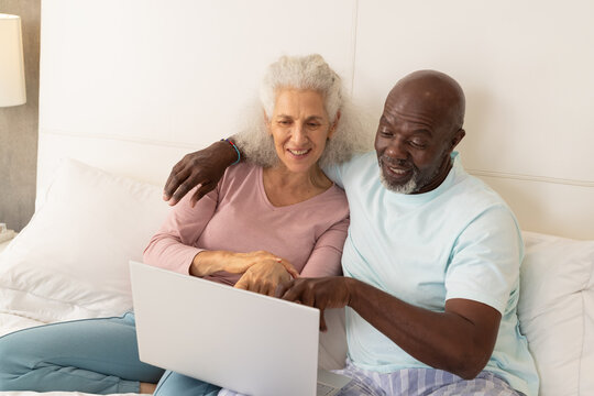 Smiling diverse senior couple leaning against white headboard in cozy bedroom, using silver laptop