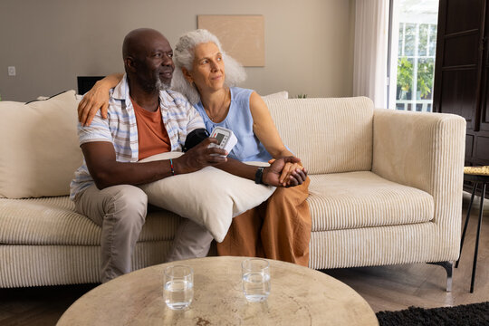 Diverse senior couple checking blood pressure in living room, with digital blood pressure monitor - Powered by Adobe