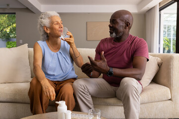 Sipping water and talking senior couple sitting on beige sofa in living room, with pill organizer