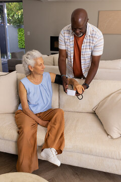 Senior African American man checking senior woman's blood pressure in living room, using BP monitor