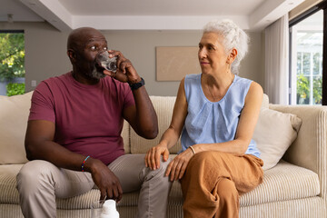 Diverse senior couple drinking water on fabric sofa in living room, with pill bottle and glasses