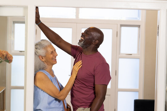 Laughing diverse senior couple leaning against door frame in hallway, with vase of peach flowers