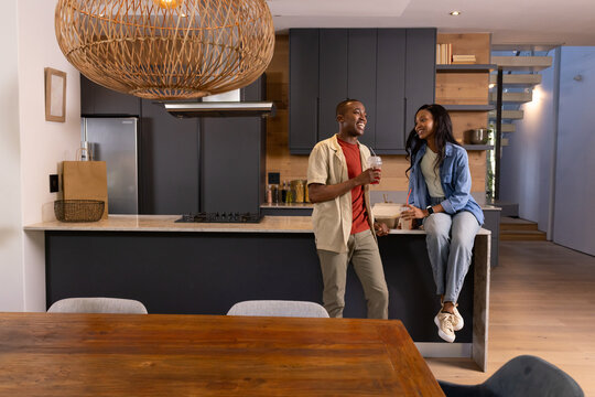 Chatting African American couple standing and sitting at kitchen island, holding bowl and clear cup