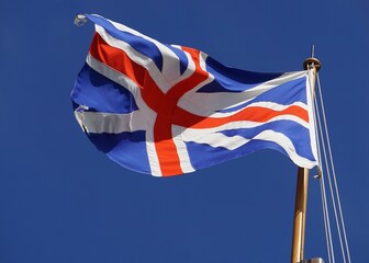 A British flag, Union Jack, patriotically waves in the wind from a historic ship mast in front of a clear blue sky