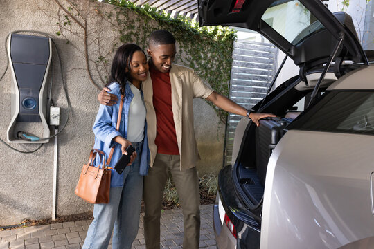 Smiling African American couple peering into silver SUV trunk at stone driveway, with EV charger