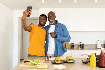 Posing African American father and son preparing burgers at kitchen island, with smartphone