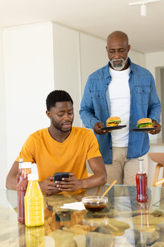 African American father offering burgers to son using smartphone at glass table in kitchen