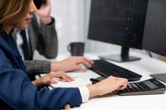 Analyzing Asian businesswoman calculating dynamic stock exchange database on computer screen with businessman partner calling customer investment online website program platform at office. Infobahn.