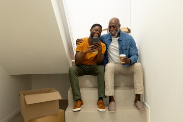 African American dad and son sitting on stairs with moving boxes, holding smartphone and coffee cup