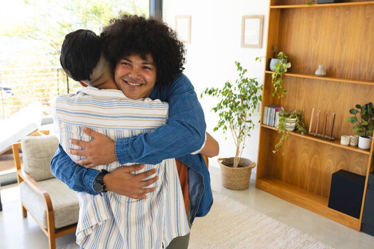 Hugging Diverse father and son standing in modern living room, with wooden bookshelf