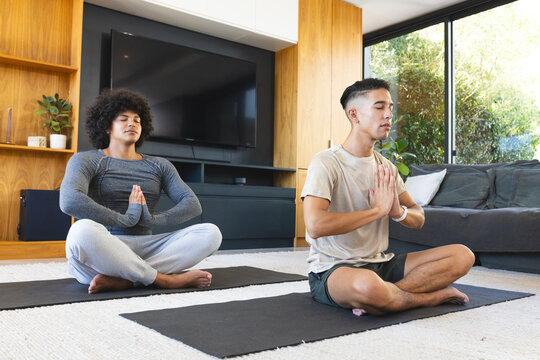 Meditating diverse male friends sitting cross-legged in modern living room, with black yoga mats - Powered by Adobe