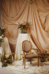 Empty classic wooden armchair surrounded by rose arrangements and soft drapery in a styled photo studio. Concept: wedding inspiration, romantic themes, elegance, or promotional content.