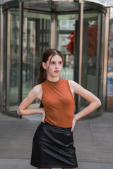 A girl with freckles in a leather skirt near a skyscraper.