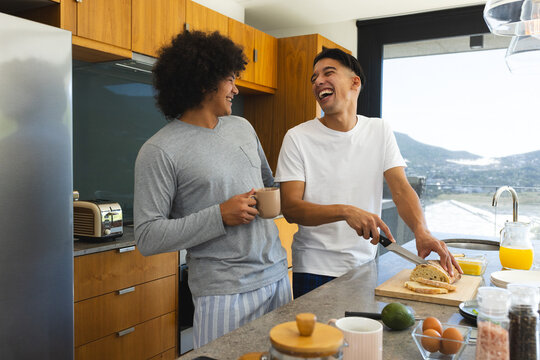 Sharing breakfast, diverse male couple slicing bread at kitchen island, with cutting board and mugs