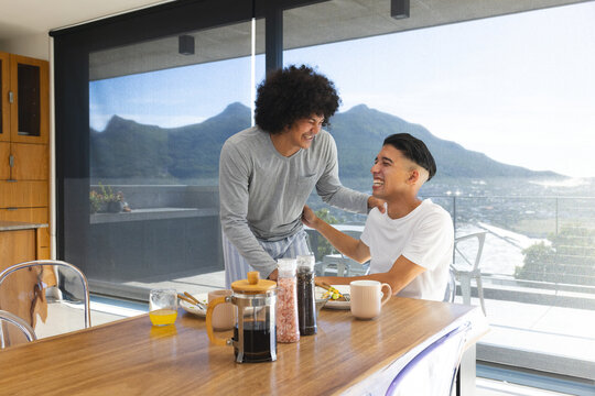 Laughing diverse male partners sharing breakfast at home dining table, with French press coffee