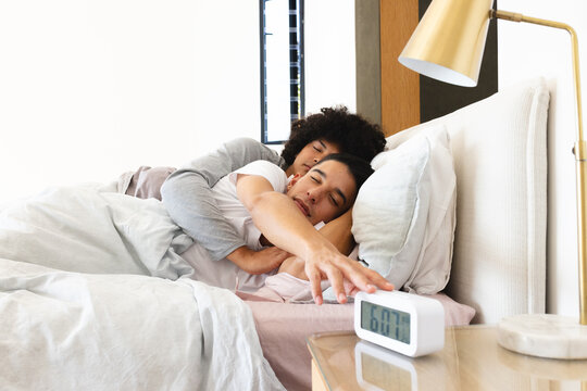 Diverse male partners reaching toward digital alarm clock in modern bedroom with white bedding