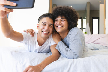 Lying Diverse male partners cuddling on bed in modern bedroom, holding tablet for selfie