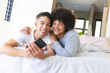 Smiling diverse male friends lying together on bed in modern bedroom, with smartphone and pillows