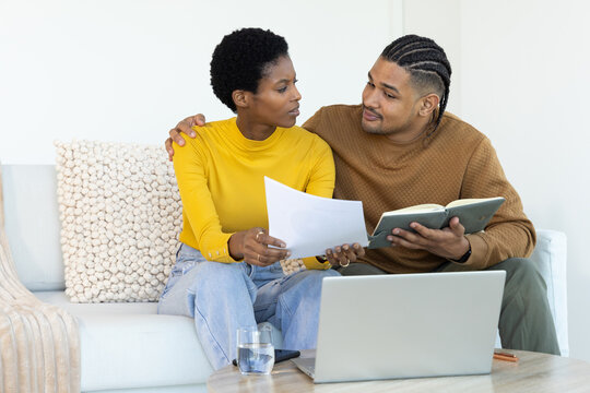 Reviewing documents Diverse couple sitting on sofa in bright living room, with laptop and notebook