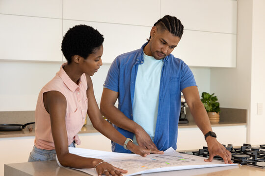 Leaning Diverse couple reviewing blueprints on kitchen island, with gas stovetop and potted plant - Powered by Adobe