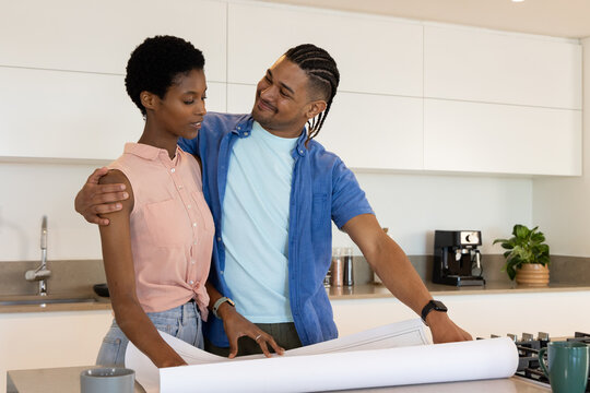 Discussing Diverse couple reviewing blueprints on kitchen island, with coffee machine - Powered by Adobe