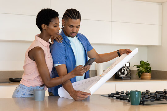 Reviewing diverse couple studying design plan on kitchen island, with smartphone and mugs