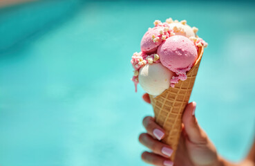 Hand holding ice cream cone with three scoops over blue water background. Tasty delicious sweet dessert. Girl enjoys vacation summer time in pool. Concept of relaxation and fun with candy.