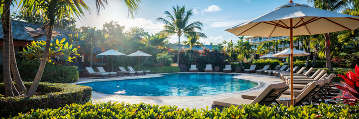 Fototapeta premium A peaceful pool area at a Caribbean resort with loungers, umbrellas, and tropical plants under a blue sky, long banner