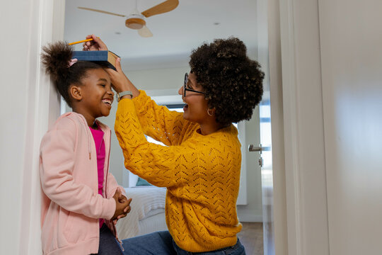 Diverse mother and daughter marking height in home doorway, with pencil and hardcover book