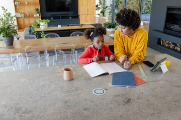 Diverse mother and daughter doing workbook tasks in modern kitchen, with pencil, copy space
