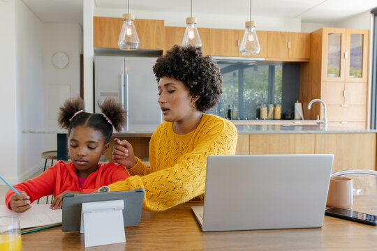 Studying Diverse mother and daughter sitting at home kitchen table, with tablet and notebooks