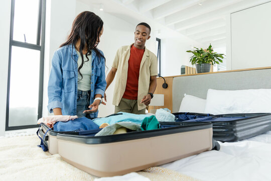 Packing African American couple sorting clothes on bed in bright bedroom, with beige suitcase - Powered by Adobe