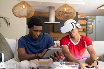 Playing games diverse male friends sitting on couch in living room, with VR headset and tablet