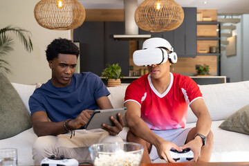 Gaming Diverse male friends tapping tablet and wearing VR headset in living room, with popcorn