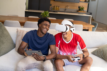 Playing VR game diverse male friends sitting on sofa at home, with VR headset and tablet