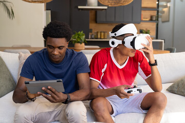 Diverse male friends using tablet and controller on sofa in living room, wearing VR headset