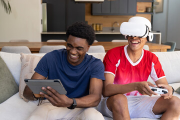 Laughing Diverse male friends using tablet and VR headset on sofa in open-plan living room