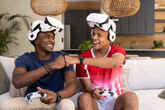 Smiling Diverse male friends fist bumping on sofa in modern living room, with VR headsets
