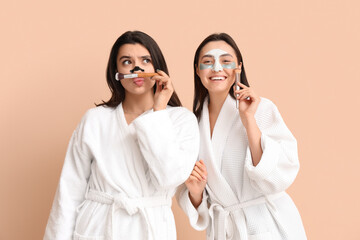 Beautiful young women in bathrobes with cosmetic masks and makeup brushes on color background